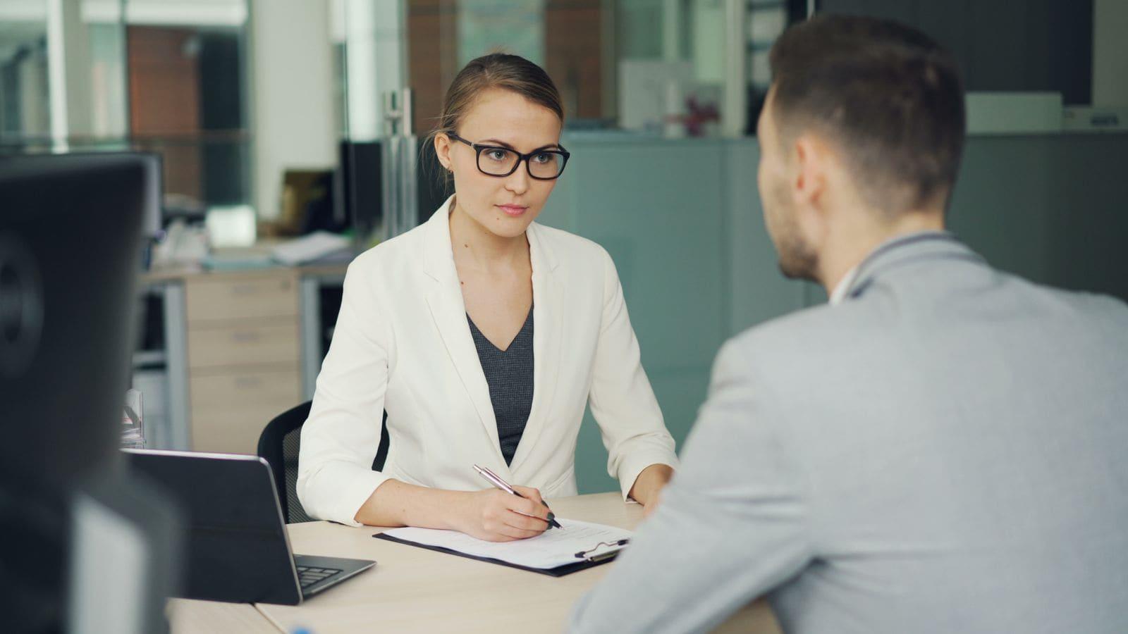 Two people talking through paperwork across a desk in a bright office